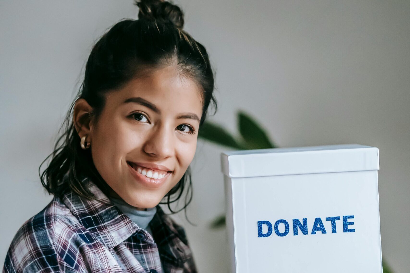 Happy ethnic lady in checkered shirt and bun looking at camera and smiling while demonstrating donation box on background with potted plant near white wall